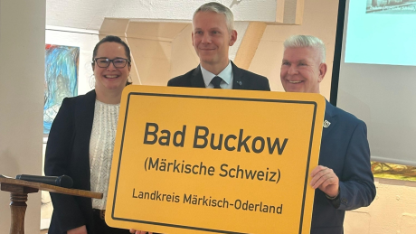 gelbes Ortsschild "Bad Buckow" mit Annett Kiesner (Tourismusleiterin des Amtes Märkische Schweiz), Marcel Kerlikofsky (Amtsdirektor) und Thomas Mix (Bürgermeister von Buckow)