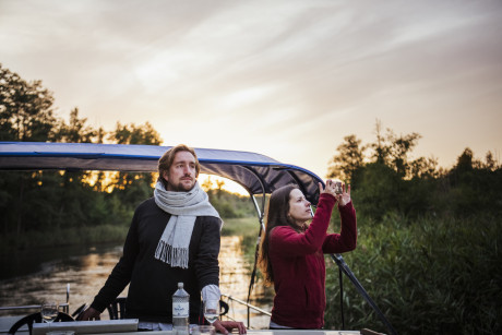 2 Personen fahren auf einem Hausboot nahe am Ufer entlang.