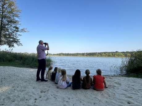 Gruppe mit Kindern sitzt am Seeufer, Erwachsener steht mit Fernglas am See und beobachtet Vögel.